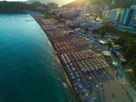 Beach with sun umbrellas, sunbeds and people relaxing in the town of Becici near Adriatic Sea against the backdrop of sunny skyの写真素材