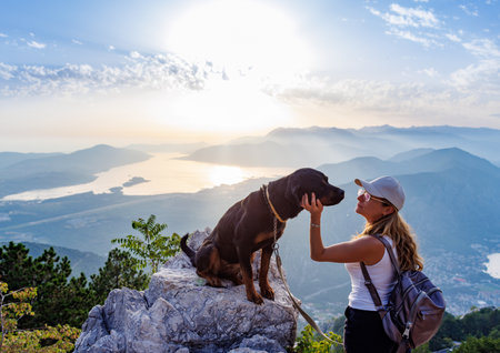 A sporty girl with a backpack stands on the edge of a mountain with a Rottweiler dogの写真素材