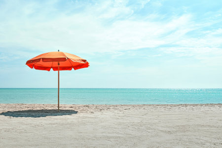 An orange beach umbrella casts a shadow on white sand, with tranquil waves gently lapping nearby.の写真素材
