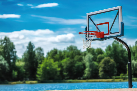 A basketball hoop stands near a calm lake, framed by greenery beneath a clear blue sky during daytime.の写真素材