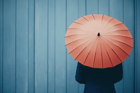 A person stands with their back to the viewer, holding an orange umbrella, in front of a blue wooden wall.の写真素材