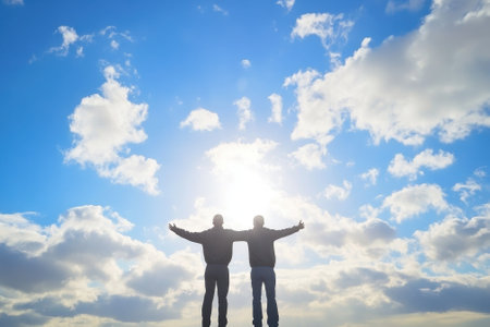 Two people stand with arms outstretched, basking in the sunlight against a beautiful sky filled with clouds.の写真素材