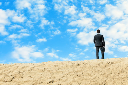 A professional man in a business suit gazes thoughtfully at the expansive blue sky while on sand.の写真素材