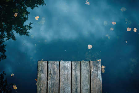 A serene view of still water mirroring the cloudy sky with a wooden dock and scattered autumn leaves.の写真素材