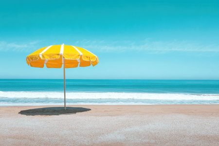 A yellow striped umbrella provides shade on a tranquil beach as calm waves gently lap at the shoreline.の写真素材