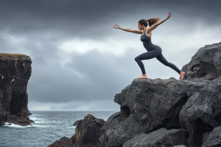 A woman performs yoga in a warrior pose on a rocky outcrop by the ocean under a cloudy sky.の素材