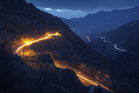 A winding road snakes through a mountainous landscape, with headlights illuminating the path as dusk sets in.の写真素材