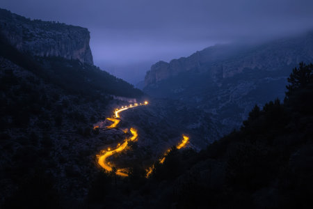 A winding road snakes through dark mountains, glowing softly from streetlights in the twilight hours.の写真素材