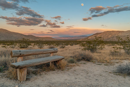 A quiet desert landscape at dusk features a wooden bench and a full moon illuminating the colorful sky.の写真素材