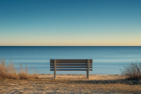 A serene bench faces tranquil waters under a clear sky at dawn, inviting peaceful contemplation by the shore.の写真素材