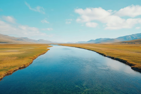A river flows through lush golden fields, surrounded by distant mountains beneath a bright blue sky.の写真素材