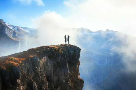 Two hikers enjoy the stunning mountain view atop a rocky cliff with clouds rolling in during daylight.の写真素材