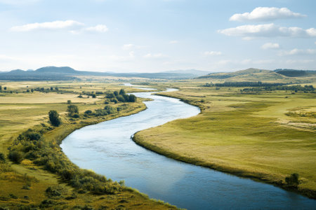 A serene river stretches through lush green fields, surrounded by distant hills under a bright sky.の写真素材