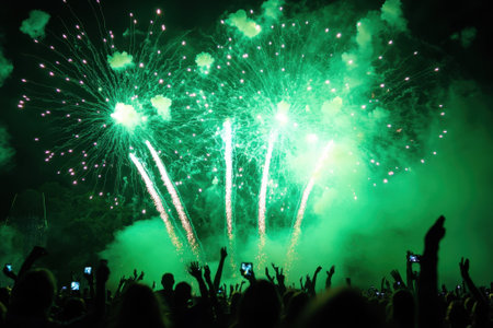 Bright green fireworks burst in the night sky, captivating a crowd as they raise their hands in excitement.の写真素材