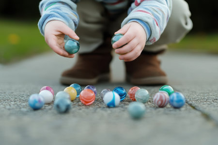 A child is crouching down to pick up marbles scattered on a paved area in a sunny park.の写真素材