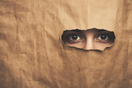 A person's curious gaze is visible through a torn hole in a sheet of brown paper, suggesting intrigue.の写真素材