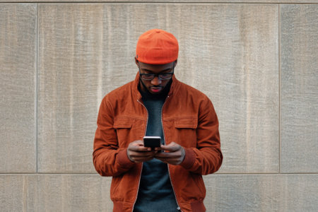 A young man wearing an orange cap checks his smartphone while standing in front of a textured wall.の写真素材