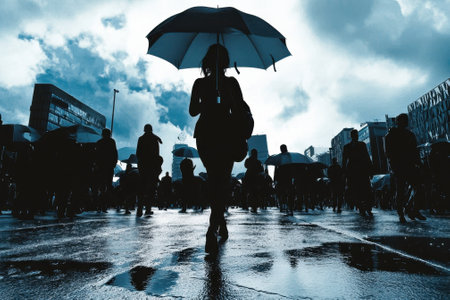 People carrying umbrellas stroll through a wet city street as dark clouds gather overhead.の写真素材