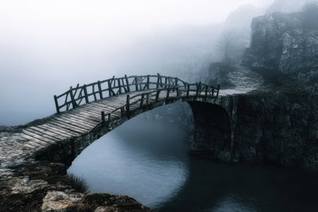 A wooden bridge arches over still water, surrounded by foggy cliffs, capturing a serene morning atmosphere.の写真素材