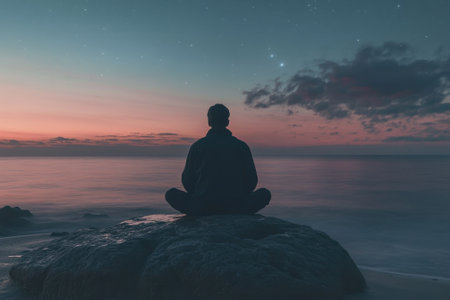 A person sits cross-legged on a rock by the water, meditating peacefully as dusk sets in and stars twinkle.の写真素材