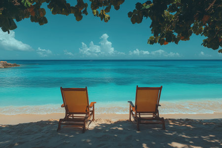 Two lounge chairs sit on a tranquil beach, inviting relaxation beneath leafy shade next to gentle waves and clear water.の写真素材