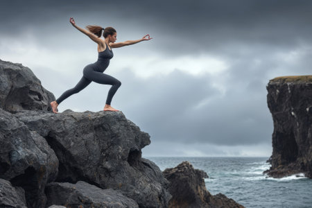 A woman performs yoga in a warrior pose on a rocky outcrop by the ocean under a cloudy sky.の写真素材