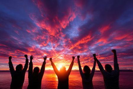 Group of friends joyfully raising their hands in celebration at the beach during a vibrant sunset.の写真素材
