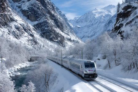 A sleek train travels through a breathtaking winter landscape with snow-covered mountains and a flowing river.の写真素材