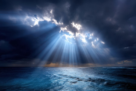 Light beams pierce through dark clouds above the serene ocean waters at twilight, creating a captivating view.の写真素材