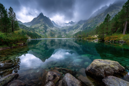 A serene mountain lake reflects peaks and cloudy skies, framed by lush greenery at the water's edge.の写真素材