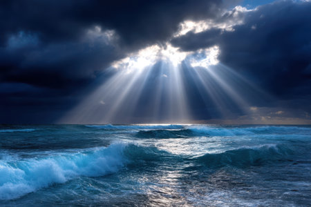 Sunlight streams down through dark clouds, illuminating rolling waves in the ocean during sunset.の写真素材