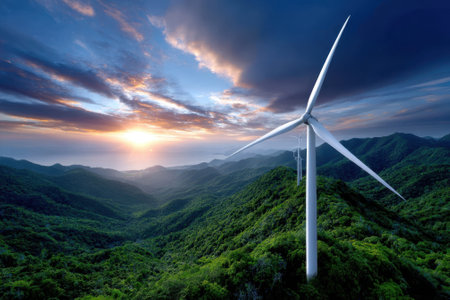 A wind turbine stands over green hills at sunset, painting the sky with vibrant colors and enhancing the landscape.の写真素材
