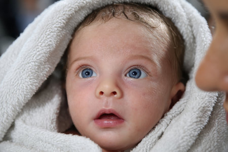 A curious baby with bright blue eyes gazes upward while being wrapped in a fluffy towel after a bath.の写真素材