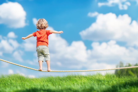 A child confidently walks on a tight rope in a grassy area under a bright blue sky with fluffy clouds.の写真素材