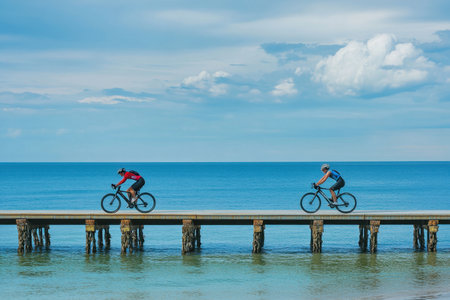 Two cyclists traverse a wooden pier while enjoying clear skies and tranquil sea views.の写真素材