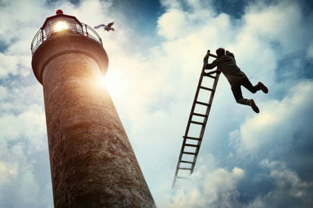A person climbs a ladder towards a tall lighthouse as a bird flies overhead in the evening light.の写真素材