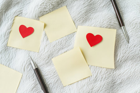 Stationery arranged with yellow sticky notes and red hearts on a cozy, soft blanket with pens nearby.の写真素材
