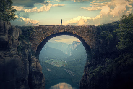 A solitary figure stands on an ancient stone bridge, gazing over a stunning valley below as the sun sets.の写真素材
