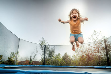 A young child leaps high on a trampoline, smiling widely under the bright sun surrounded by greenery.の写真素材