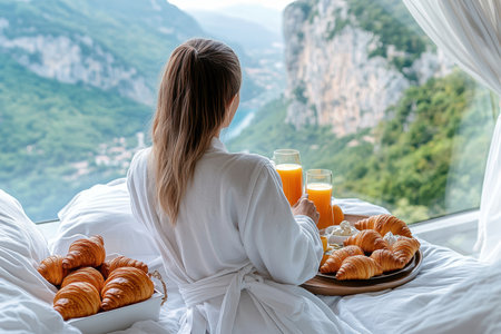 Relaxing in a luxurious robe, a woman savors fresh croissants and juice by a panoramic window.の写真素材