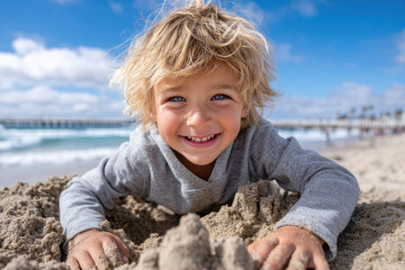 Young boy smiles while digging in the sand on a lively beach, enjoying his playful moment by the ocean.の素材