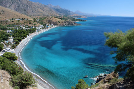 Expansive view of a serene beach with turquoise waters, rocky hills, and lush green trees under a clear blue sky.の写真素材