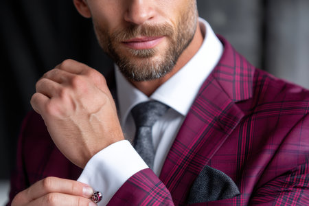 Man with a beard fine-tunes cufflinks in a burgundy plaid suit, showing elegance and style in a chic interior.の素材