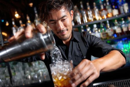A skilled bartender prepares a cocktail by pouring ingredients over ice, showcasing his craft in a lively bar.の素材
