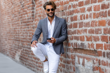 Stylish man leans against a brick wall, dressed in a blazer and white jeans, checking his phone.の素材