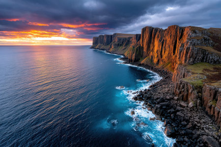 Rocky cliffs meet a vibrant ocean under a dramatic sunset skyの写真素材