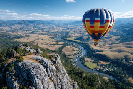 A colorful hot air balloon floats gracefully over a lush valley and winding river under a clear blue sky.の写真素材