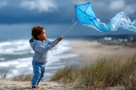 A young child joyfully flies a blue kite on a coastal beach while waves crash nearby under overcast skies.の写真素材