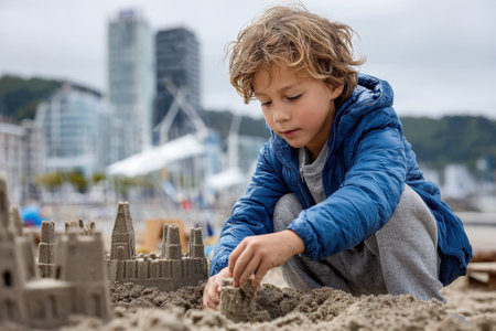 A boy skillfully molds a sandcastle on the beach while city buildings rise in the background.の写真素材
