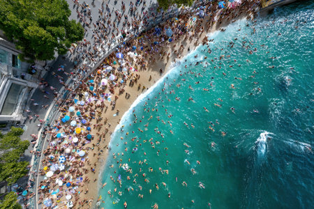 Excited visitors enjoy sunbathing and swimming at a popular beach, surrounded by vibrant umbrellas and waves.の素材